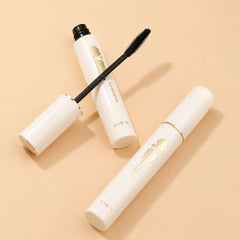 BCM O KOL white cosmetic mascara tubes with gold accents and a black applicator wand, arranged on a warm beige background.